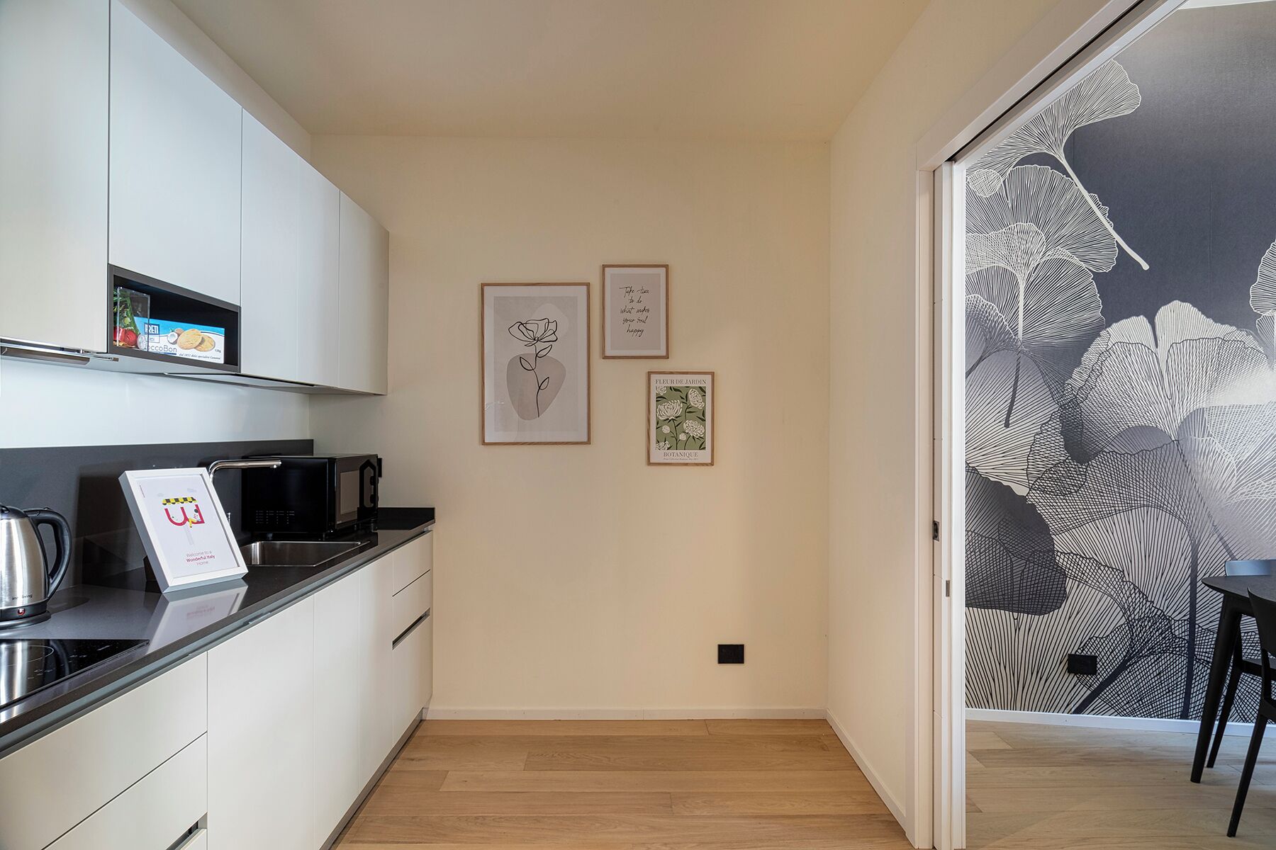 Een moderne keuken met witte kasten, een zwart aanrecht en een waterkoker. Er hangen drie ingelijste foto's aan de muur.