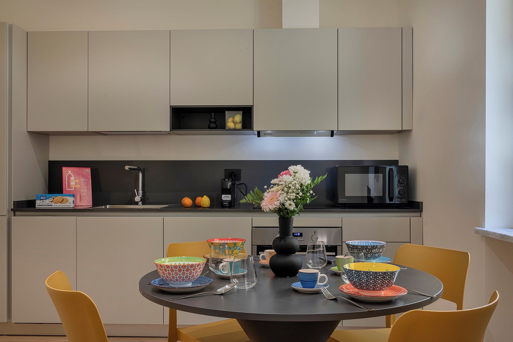 Une cuisine moderne avec un plan de travail noir, un four à micro-ondes et un évier. On peut voir des bols colorés et un vase rempli de fleurs sur la table.