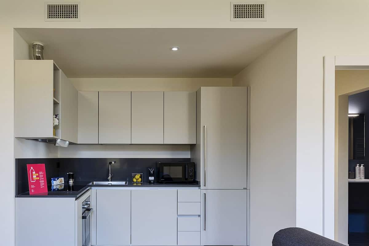 Une cuisine compacte avec des meubles blancs, un plan de travail noir et un four à micro-ondes. On y trouve un petit évier avec un robinet, ainsi qu’une boîte rouge posée sur le plan de travail.