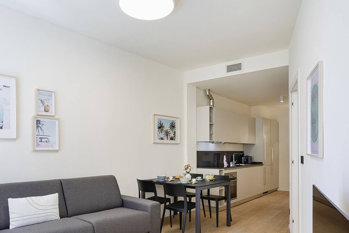 Un espace de vie compact comprenant un canapé gris, une table à manger avec des chaises, un coin cuisine et des œuvres d’art encadrées accrochées aux murs.