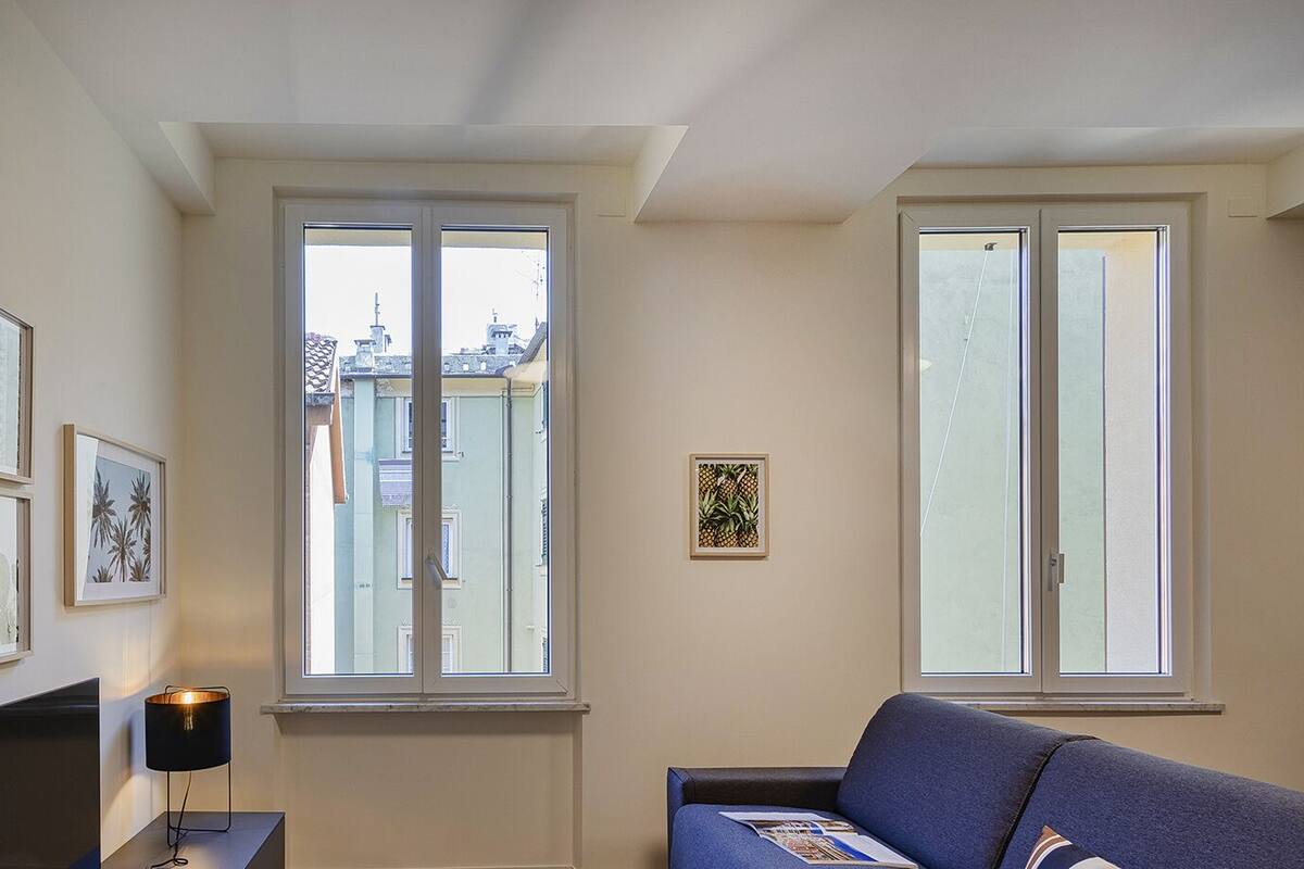 Un salon comprenant un canapé, un téléviseur, des œuvres d’art encadrées et deux fenêtres donnant sur des immeubles.