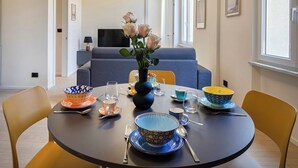 A dining area with a round table set for four, featuring blue and orange patterned bowls, a vase with flowers, and a modern sofa in the background. - Riviera Flavour Apartments II by Wonderful Italy (Santa Margherita Ligure)