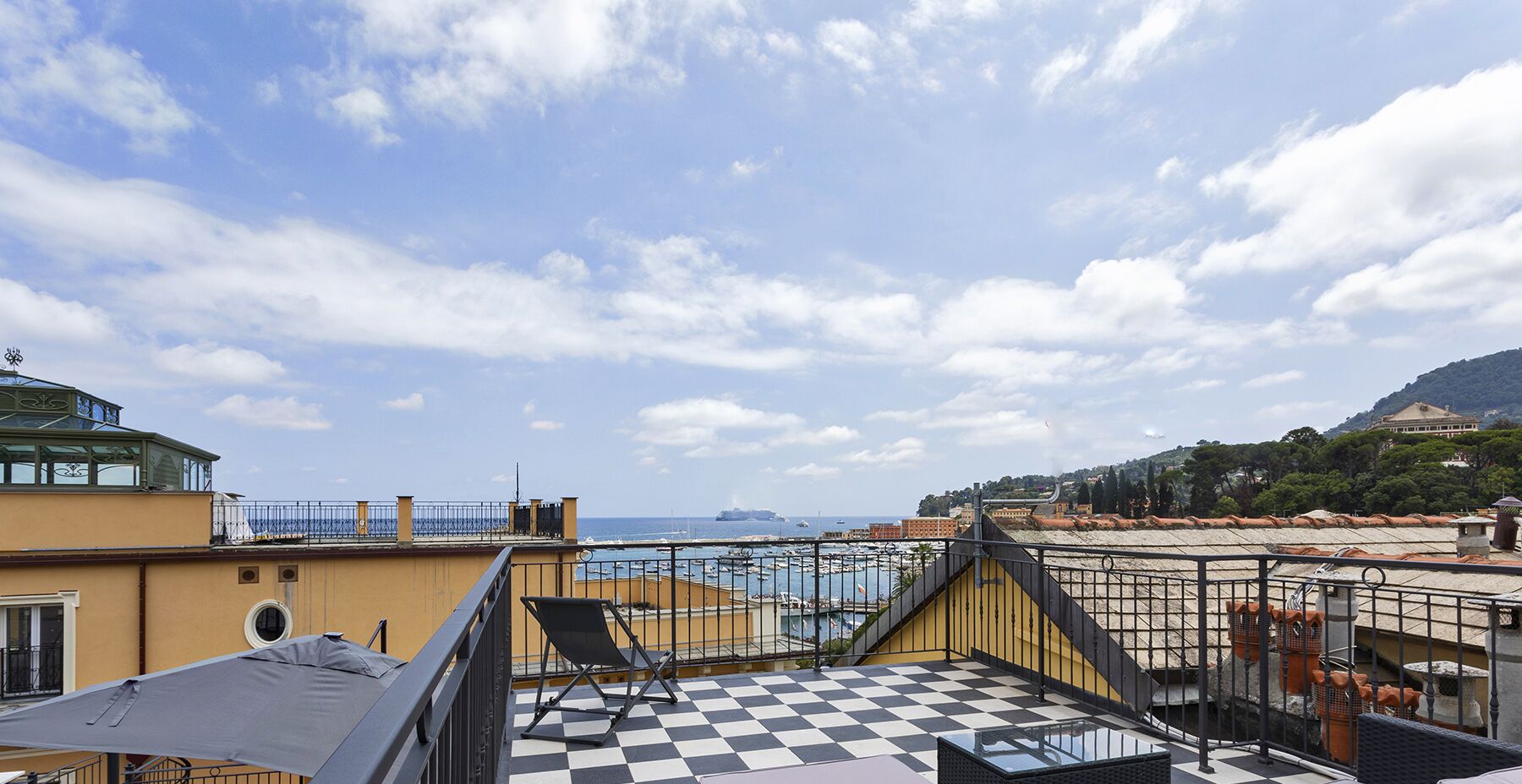 A rooftop terrace with a checkered floor, a view of the sea, and a distant cityscape.
