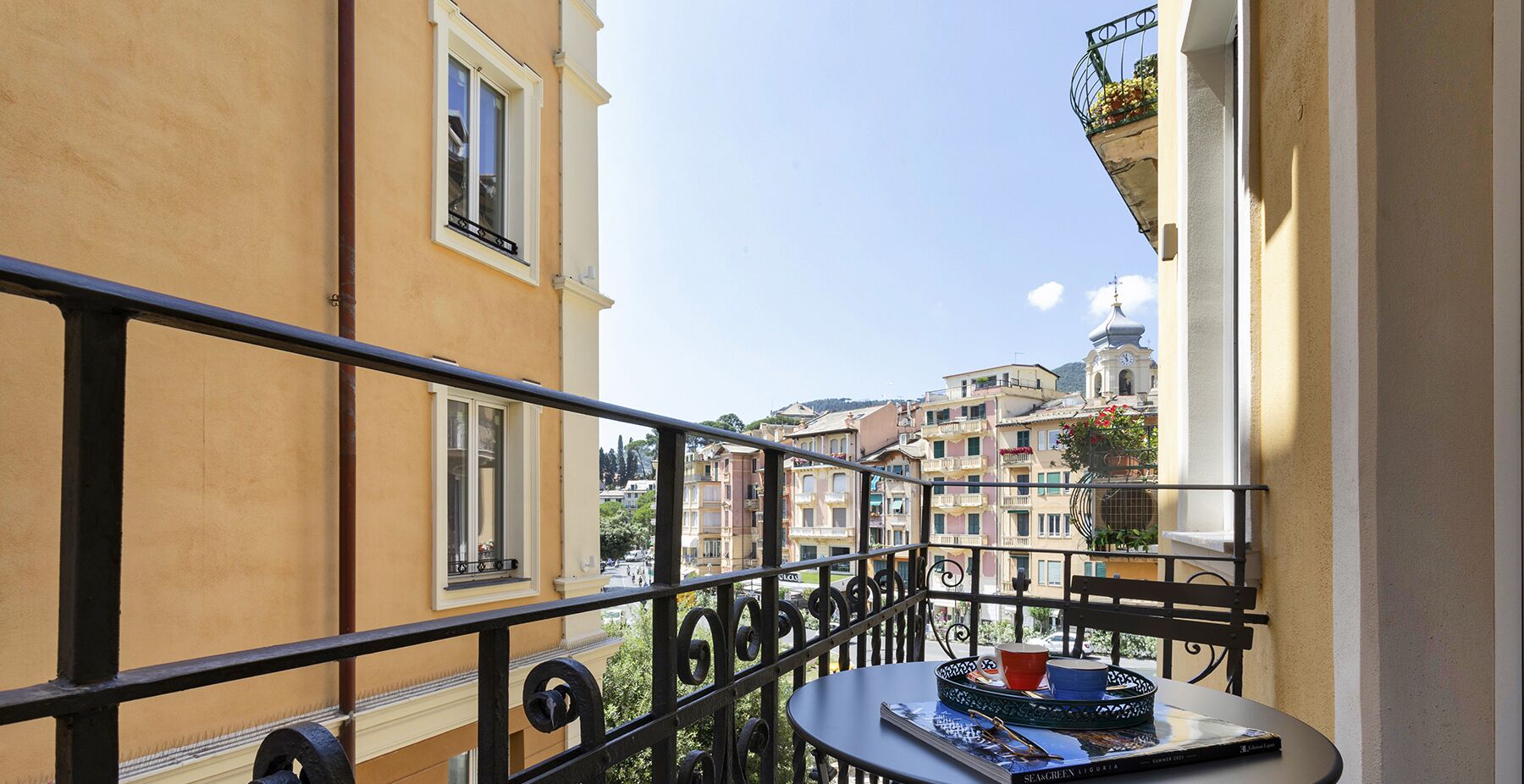 Un balcon avec une table, une chaise et une vue sur une ville avec ses bâtiments et le clocher d’une église.