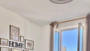 Quarto moderno com mesa de vidro e cadeiras brancas, um letreiro "HOME" na parede e vista para o mar por uma janela aberta.