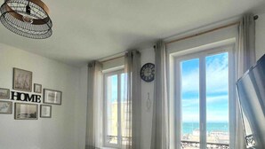 Quarto com mesa de jantar, sofá, ventilador de teto, relógio e vista para o mar através de grandes janelas.