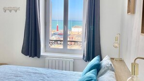 Quarto com cama, janela com cortinas, radiador, mesa de cabeceira com abajur e vista para a praia.