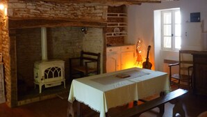 Une pièce rustique avec une cheminée en pierre, des poutres en bois, une table recouverte d’une nappe et une guitare.