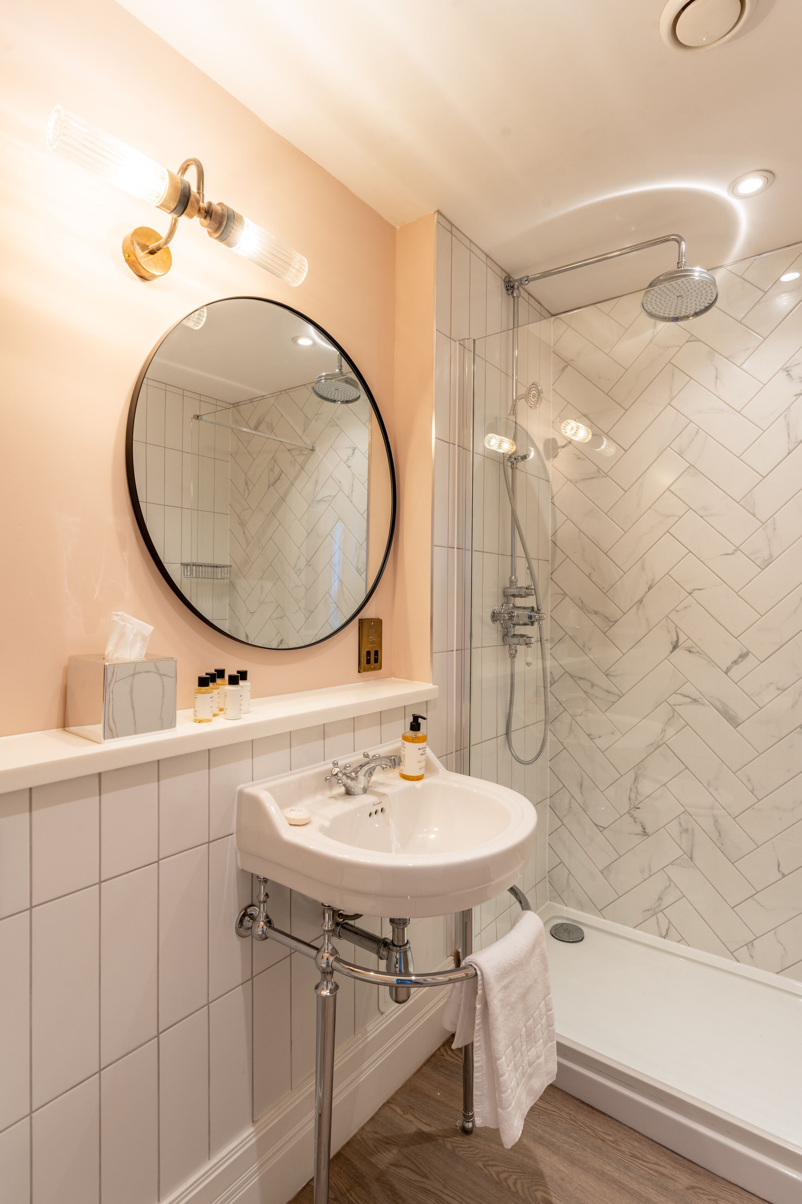 A bathroom with a round mirror, a sink, a shower, and a towel hanging on the door.