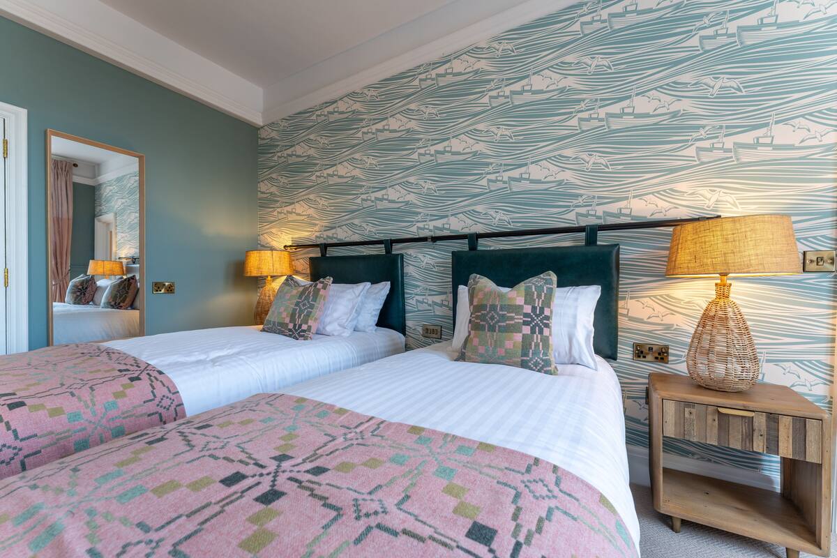 A double bed with patterned bedding, a nightstand with a lamp, and a wall with a nautical-themed wallpaper.