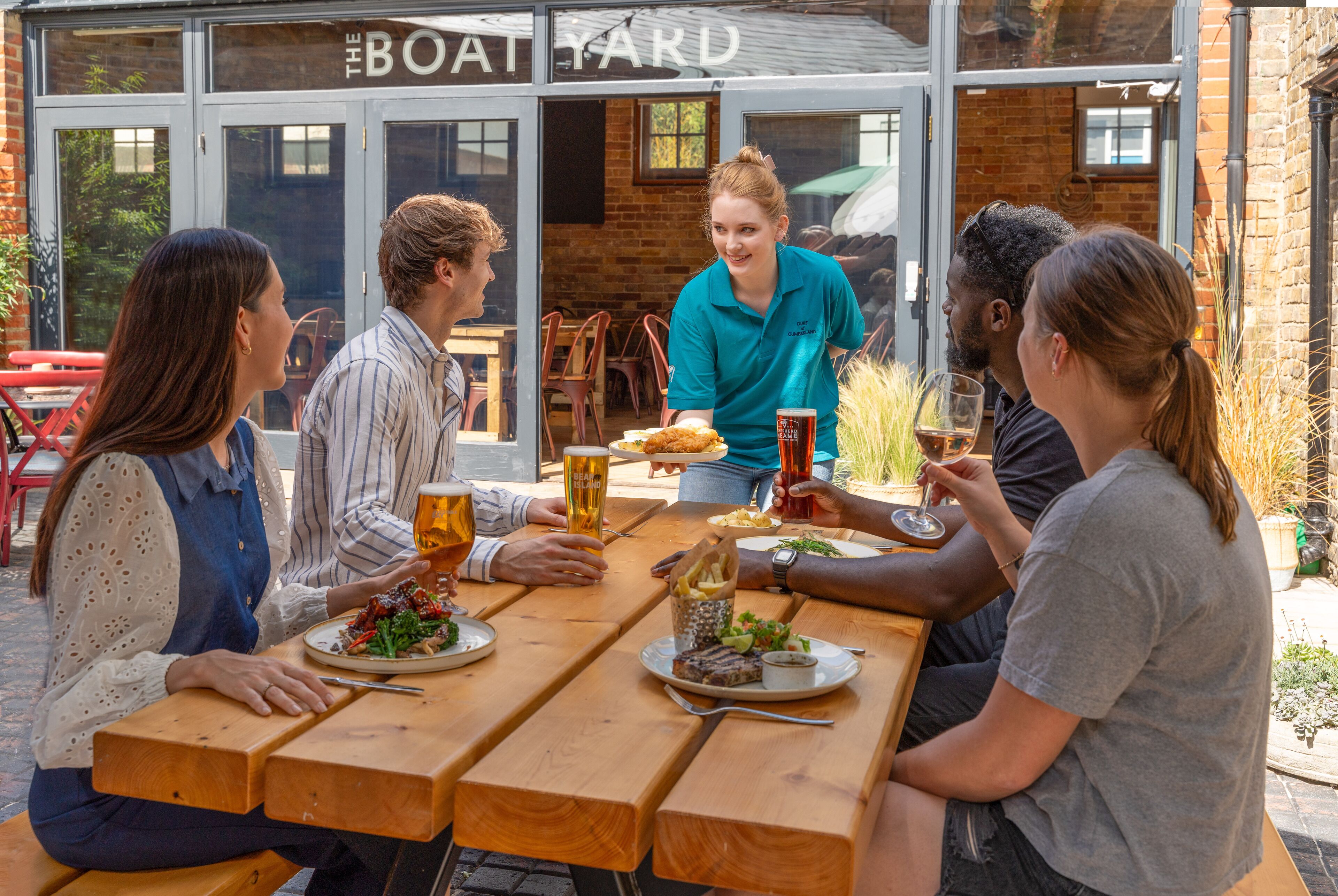 Un grupo de personas comiendo y bebiendo en una mesa al aire libre frente al restaurante "The Boat Yard".