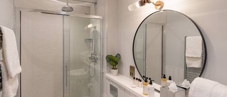 A modern bathroom with a glass-enclosed shower, a round mirror, and a vanity with toiletries.