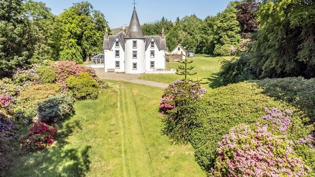 Un grand bâtiment blanc à plusieurs étages, doté d’un toit en forme de flèche et d’une tour centrale, entouré de jardins et d’arbres parfaitement entretenus.