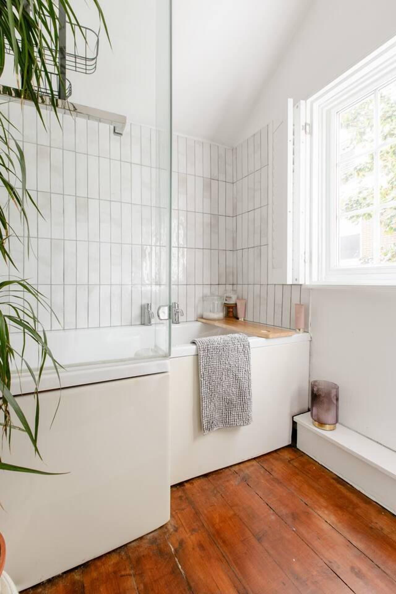 Un baño moderno con cabina de ducha de vidrio, paredes de azulejos blancos, suelo de madera y una ventana que proporciona luz natural.