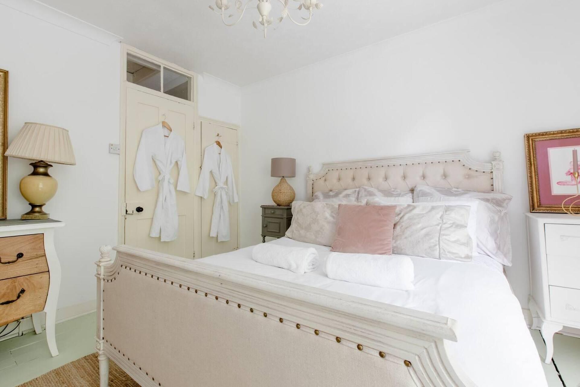 A white bedroom with a four-poster bed, a dresser, a nightstand, and a wardrobe with two white robes hanging.