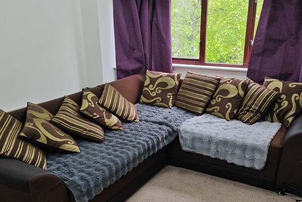 A corner sofa with patterned cushions, a blue throw blanket, and a view of green foliage through a window with purple curtains. - 1-bed Apartment in Croydon -free Parking, Wifi (Croydon)