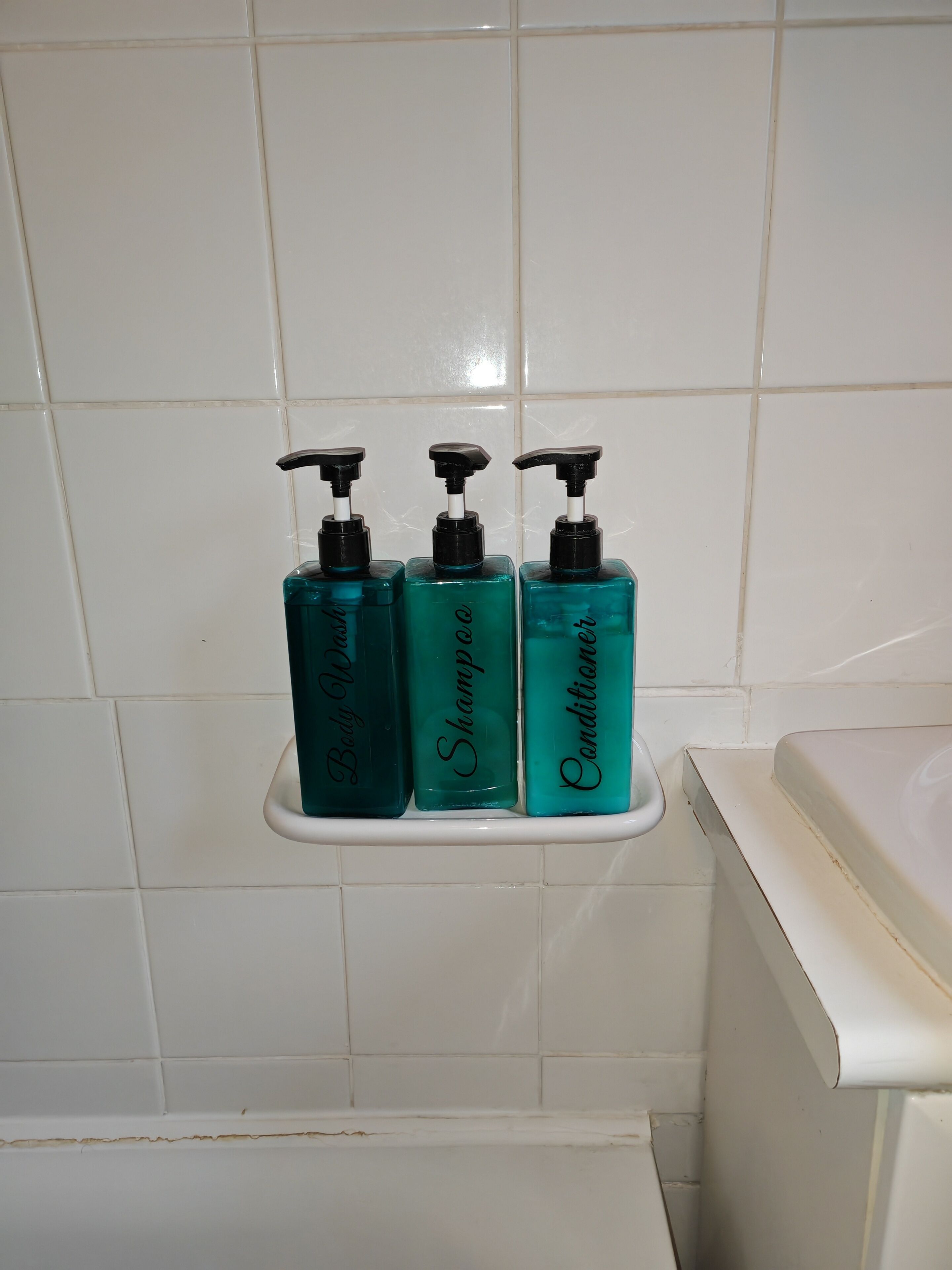 A bathroom with white tiled walls and a white bathtub. There are three bottles of liquid soap on a white shelf.