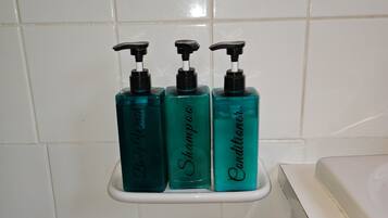 A bathroom with white tiled walls and a white bathtub. There are three bottles of liquid soap on a white shelf.