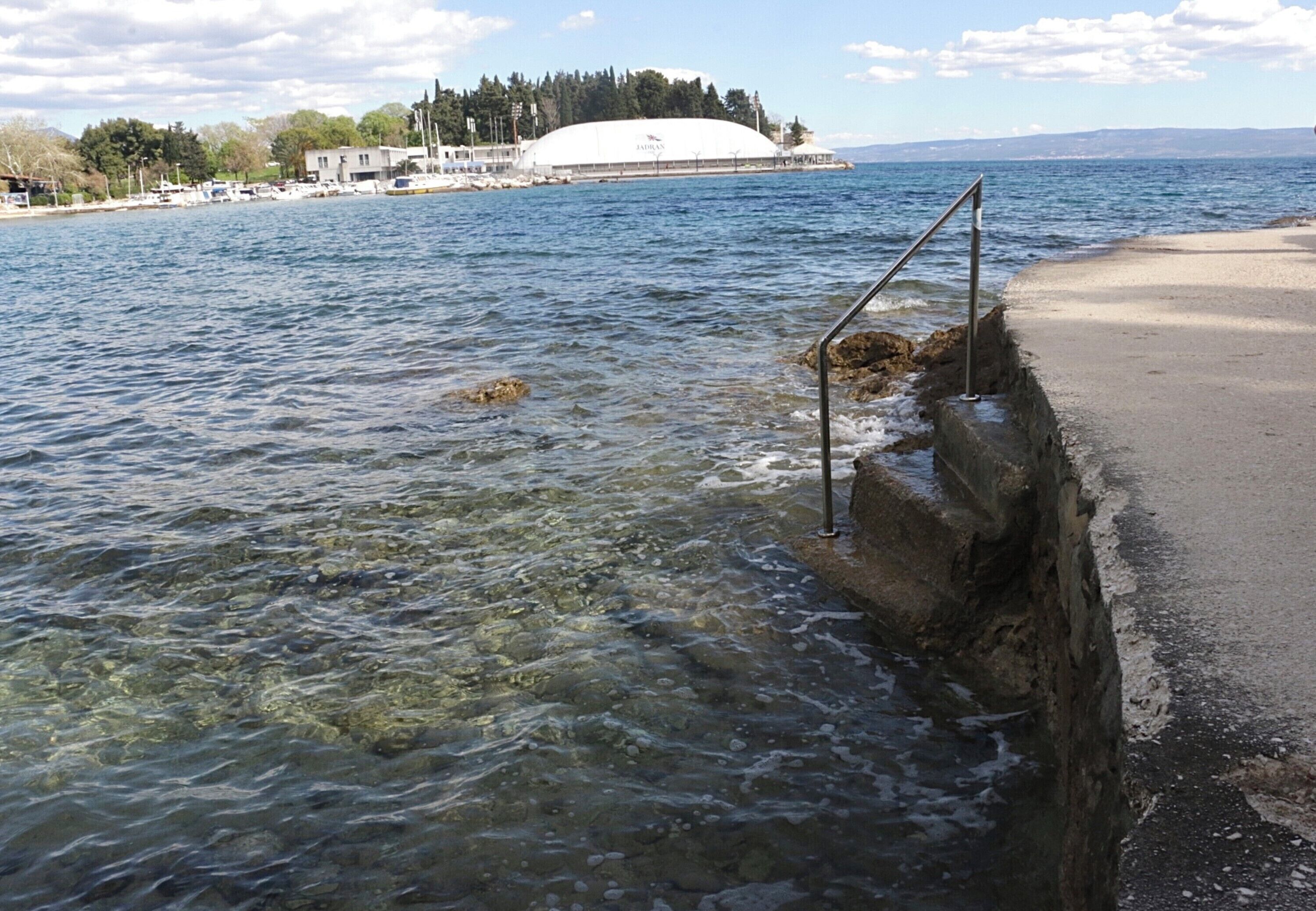 A waterfront view with clear water, a concrete barrier with a metal handrail, and a large white dome structure in the background.
