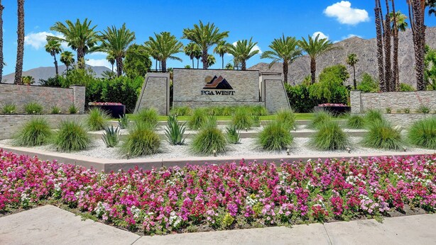 A landscaped entrance with a sign reading "PGA WEST" surrounded by palm trees and flowering plants. - Brand New at PGA West, views, community pool, sleeps 10 #254654 (La Quinta)
