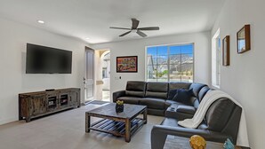 A modern living room with a large sectional sofa, a coffee table, a TV mounted on the wall, and a ceiling fan. - Brand New at PGA West, views, community pool, sleeps 10 #254654 (La Quinta)