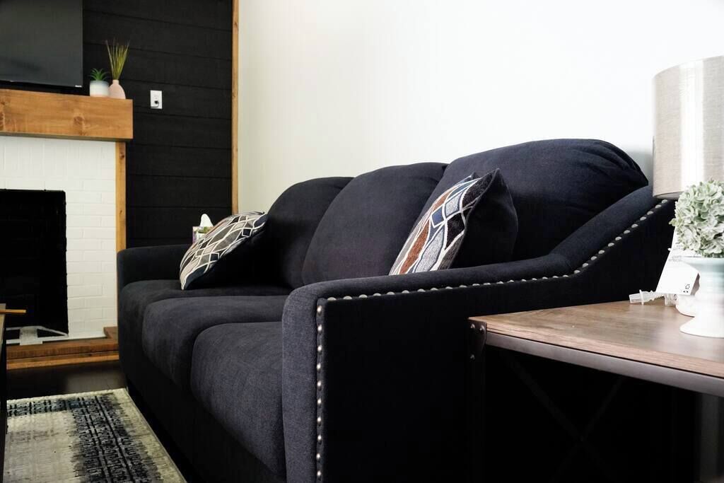 Een zwarte bank met decoratieve kussens, een houten tafel en een witte vaas met een plant.