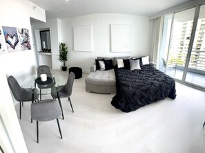 A modern bedroom with a large bed, a sofa, a dining table with chairs, and a balcony with a view of a high-rise building.