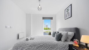 A modern bedroom with a large bed, a nightstand, a lamp, and a view of buildings through a window.