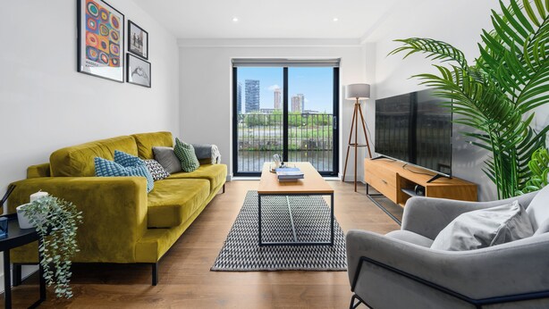 A modern living room with a yellow sofa, a grey armchair, a wooden coffee table, and a large TV. There is a black and white rug, a potted plant, and artwork on the wall.