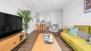 A modern living room with a flat-screen TV, a yellow sofa, a wooden coffee table, and a dining area in the background.