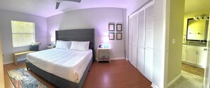 Ein Schlafzimmer mit Bett, Nachttischen, einer Lampe, einem Fenster mit Jalousien, einer Tür mit Klappläden und einem Badezimmer mit Spiegel und Waschbecken.