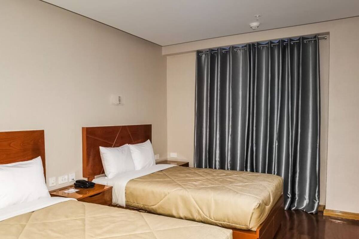 Habitación de hotel con dos camas, cabecero de madera, un ventanal grande con cortinas y una mesita de noche con teléfono.