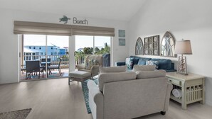 A beach-themed living room with a sofa, armchairs, a dining area, and a balcony with a view of buildings. - Close to Beach - In-Home Peloton Retreat + Free Attraction Tickets! (Miramar Beach)