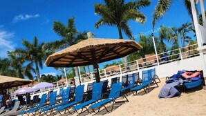 A beach with rows of blue lounge chairs and thatched umbrellas, palm trees in the background, and a white building with a balcony. - Free Beach&pool Access -vaes Luxury Escape (Discovery Bay)
