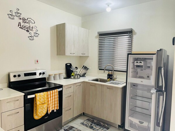 Una cocina moderna con estufa, horno, fregadero y refrigerador. Hay armarios, una ventana con persianas y un elemento decorativo en la pared.