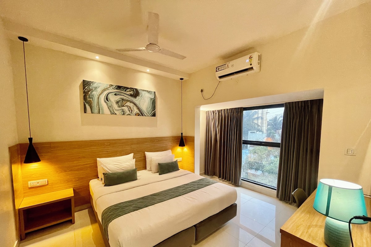 A hotel room with a bed, bedside lamps, a ceiling fan, an air conditioning unit, a window with curtains, and a painting on the wall.
