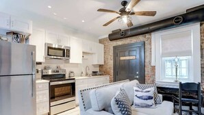 A modern kitchen with white cabinets, stainless steel appliances, and a brick accent wall. - Cozy Historic 1790 Guest House Steps to River St (Savannah)