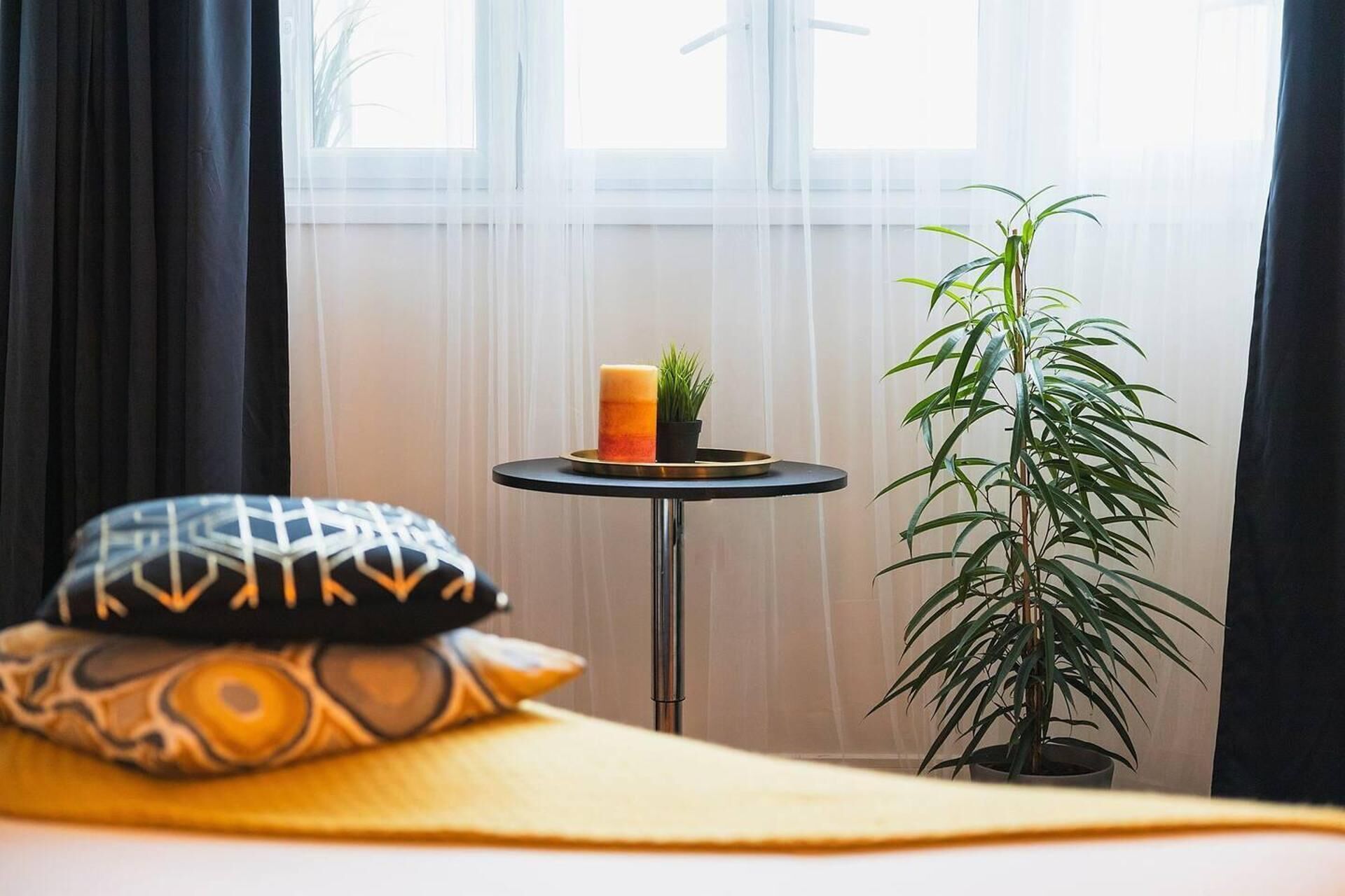 A bed with a yellow blanket, a patterned pillow, a small table with a candle and a plant, and a tall green plant beside the window.