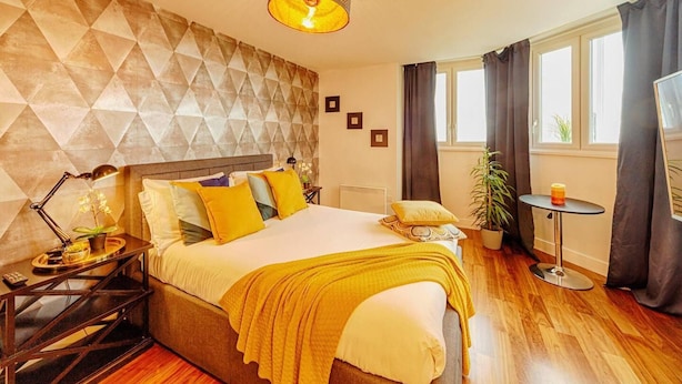 Una camera d'albergo moderna con una testiera geometrica, un letto grande con molti cuscini, un comodino con una lampada, un tavolino rotondo con una candela e una pianta in vaso.
