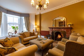 A cozy living room with a fireplace, comfortable seating, and a large window with curtains. - Craigroyston House & Lodge (Pitlochry)