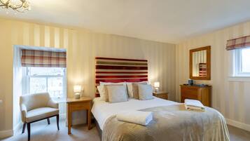 A hotel room with a large bed, a bedside table, a chair, a window with blinds, a mirror, and a chandelier.
