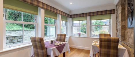 A dining area with a table set for two, flanked by plaid chairs, large windows with views of greenery, and a stone wall.