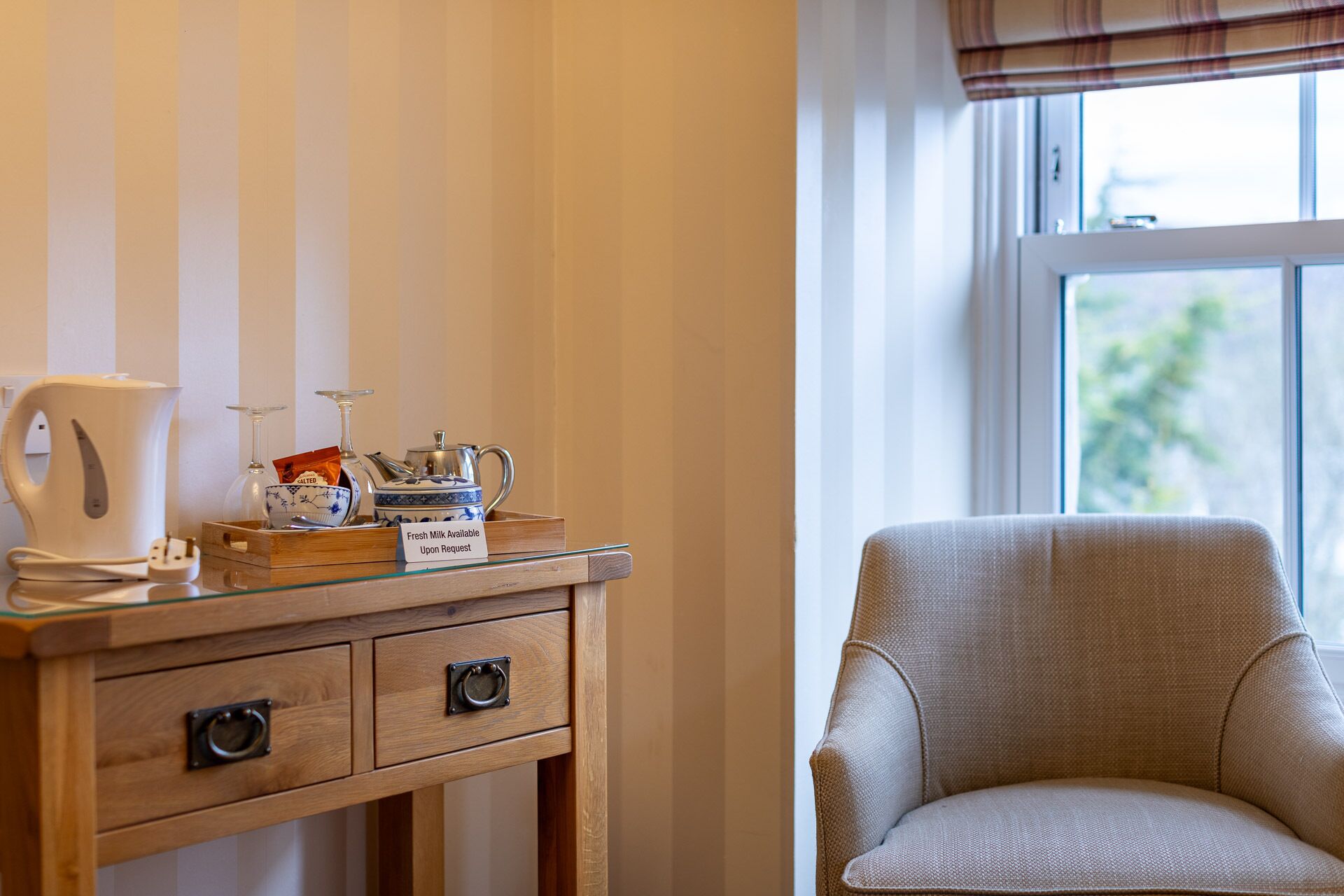A wooden side table with a kettle, teapot, and a sign indicating fresh milk availability. A chair is positioned near a window with a view of greenery.
