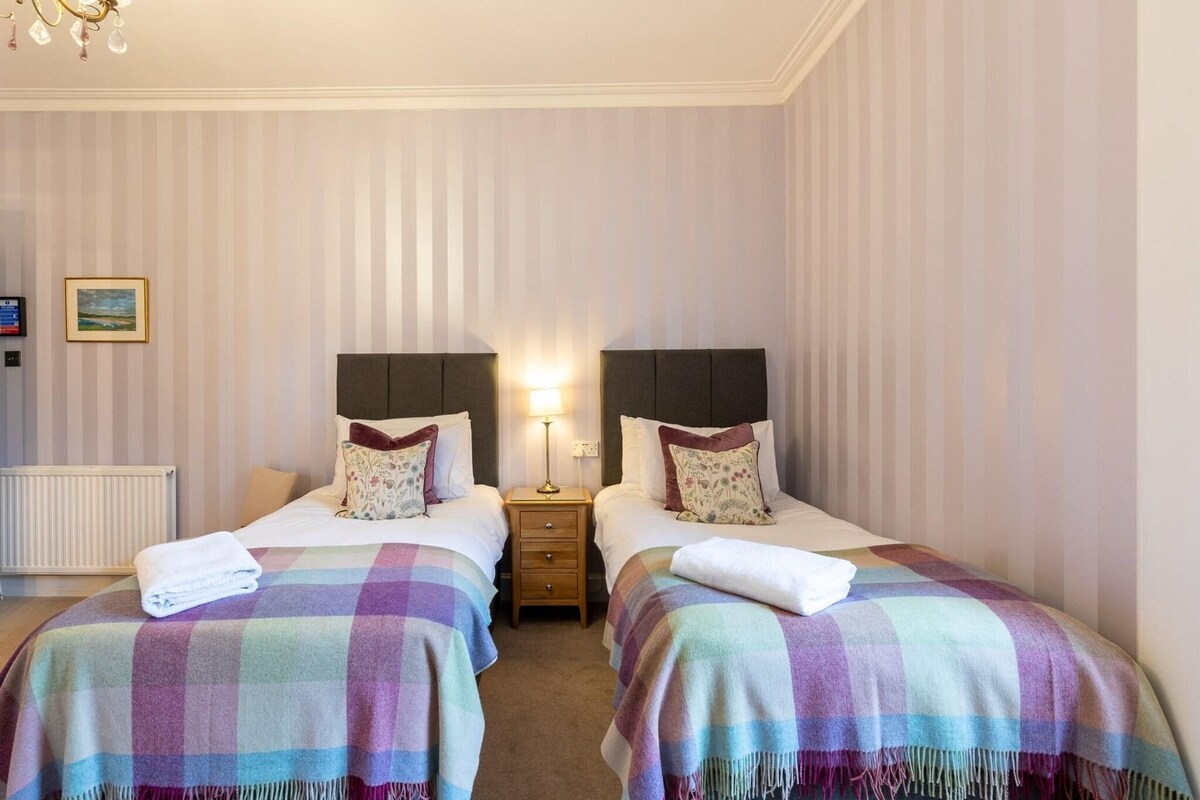 A hotel room with two beds, striped wallpaper, a radiator, a nightstand with a lamp, and a framed picture on the wall.