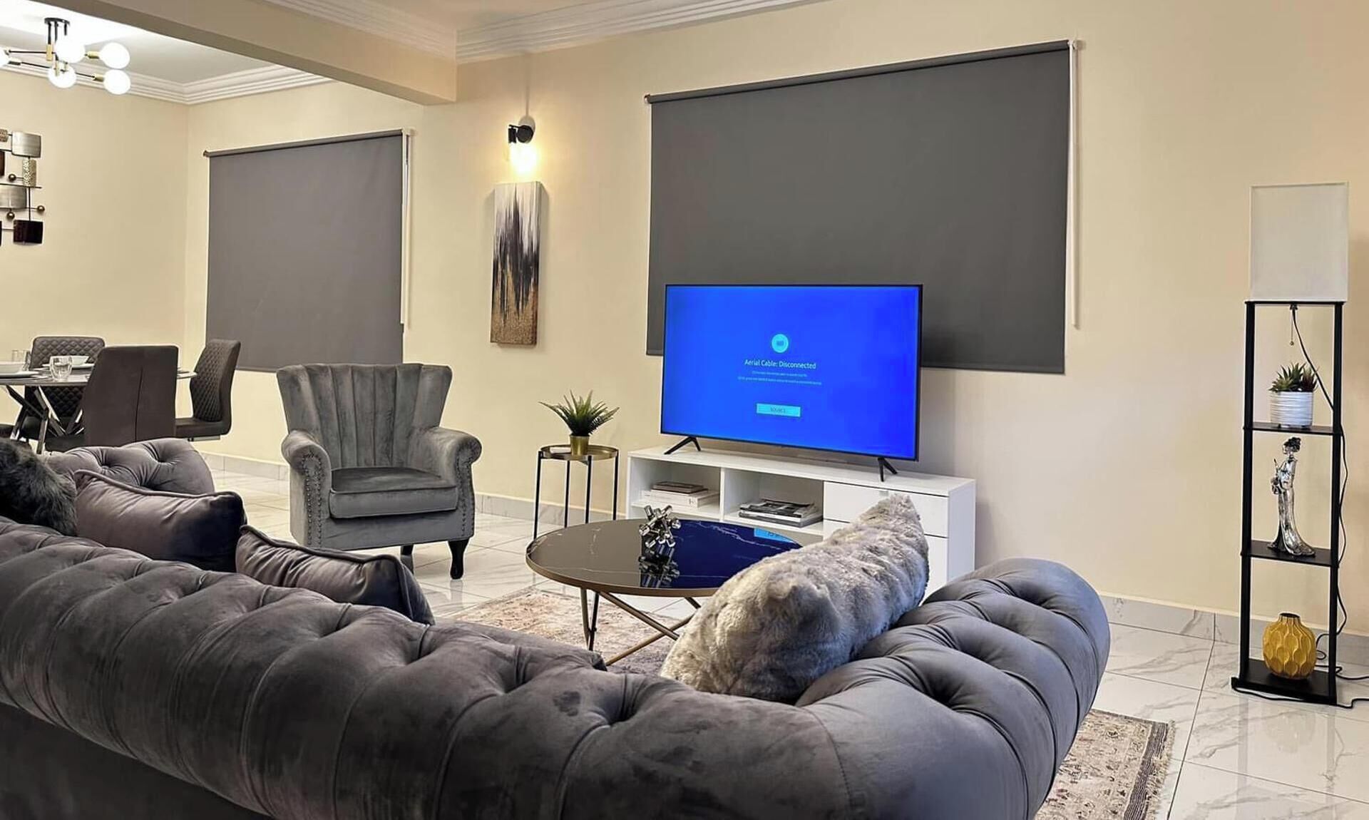 A modern living room with a large flat-screen TV, grey upholstered sofa, and a dining area with a table and chairs.