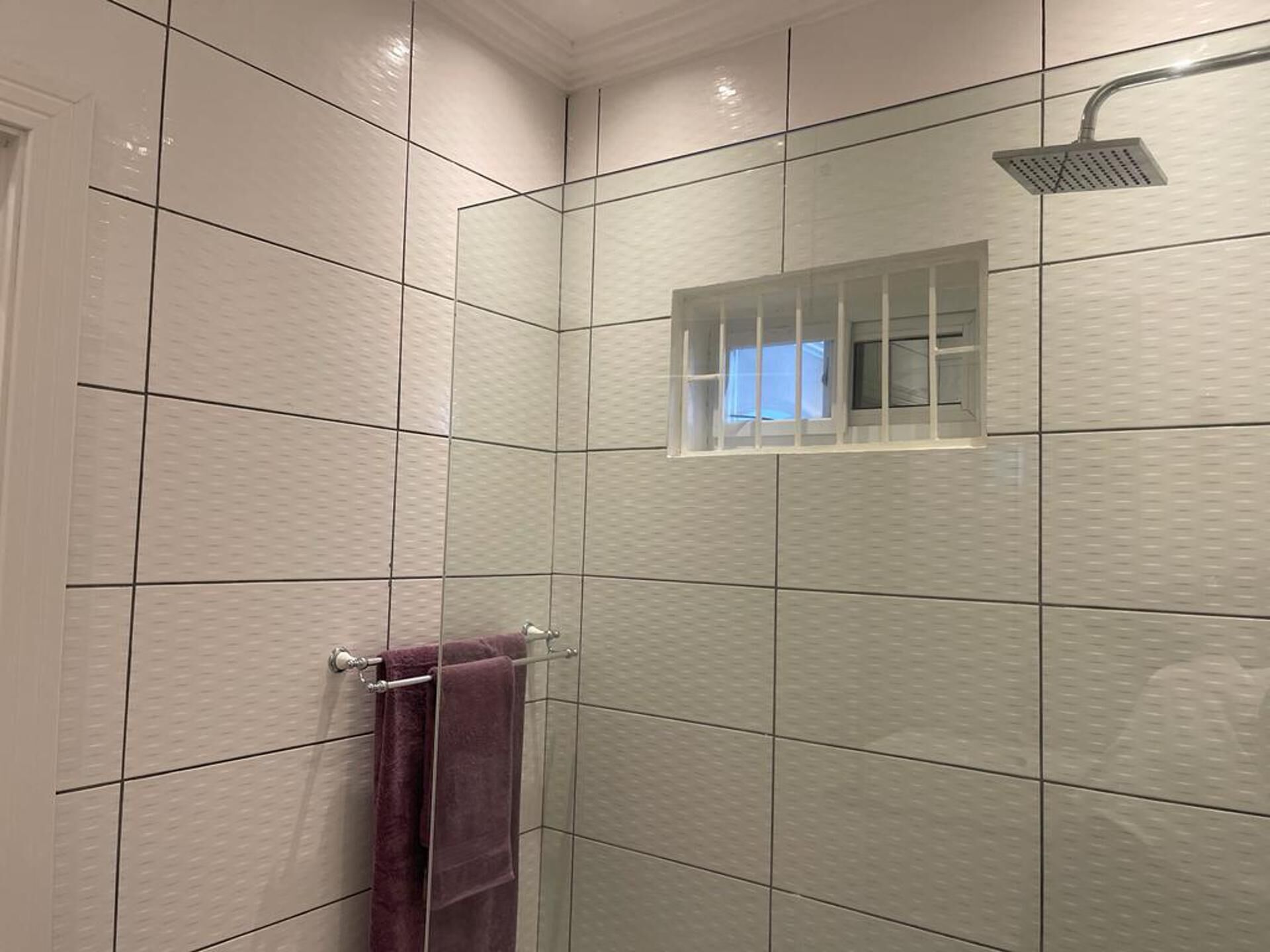 Une salle de bain avec des murs carrelés de carreaux blancs, une douche dotée d’une paroi en verre et une fenêtre visible.