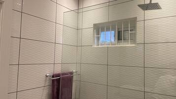 Une salle de bain avec des murs carrelés de carreaux blancs, une douche dotée d’une paroi en verre et une fenêtre visible.