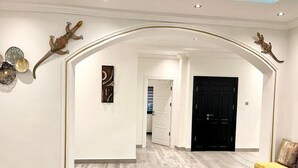 A hallway with a wooden floor, white walls, and a black door at the end. Decorative wall art and a yellow chair are also visible.