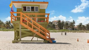 A lifeguard tower with a wooden staircase, a flag, and lifeguard chairs on a sandy beach.