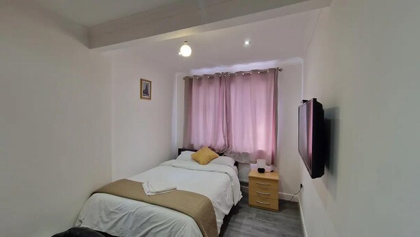 A bedroom with a bed, a bedside table with a lamp, a TV mounted on the wall, and a window with curtains.